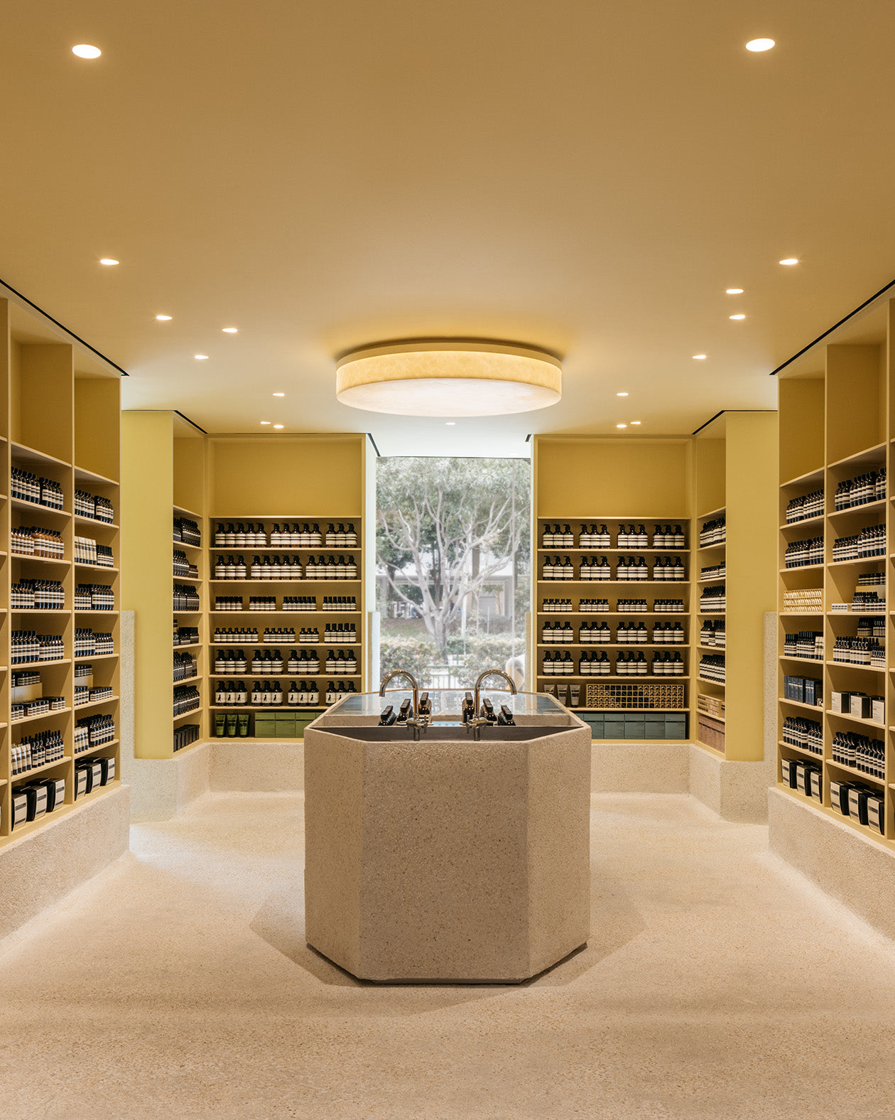 A large hand-cast pebble wash monolith in the middle of the Tampines store with two basins next to it and the smooth shelves on the surrounding walls are lined with Aesop products.