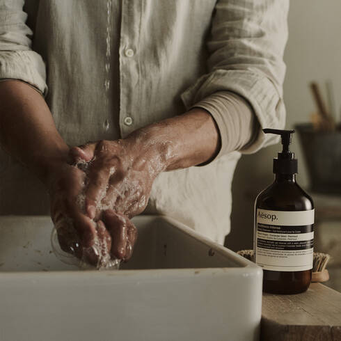 Hands being washed at a sink beside an Aesop Antithesis Intense Body Cleanser pump bottle.